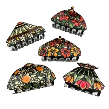 Five decorative claw hair clips arranged on a white surface, each featuring a different colorful floral and fruit pattern with black claw bases. A partial book and wooden object are visible in the background.