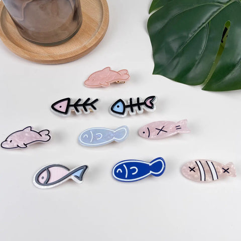 A set of fish-shaped hair clips arranged on a white surface, with a wooden tray edge and a green leaf in the background. The clips feature various fish designs in pastel pink, light blue, dark blue, and black-and-white skeletal patterns.