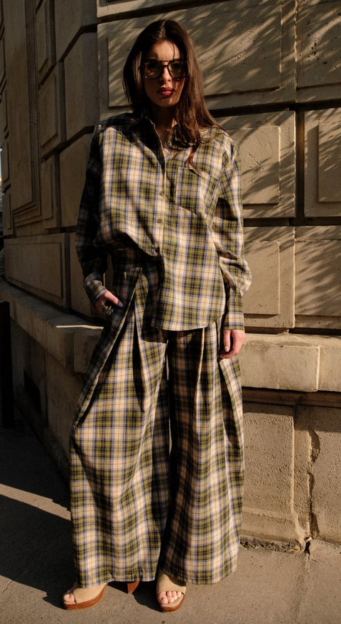 A woman wearing a matching plaid button-up shirt and wide-leg pants set, styled with sunglasses and flat sandals, standing outdoors against a stone wall.