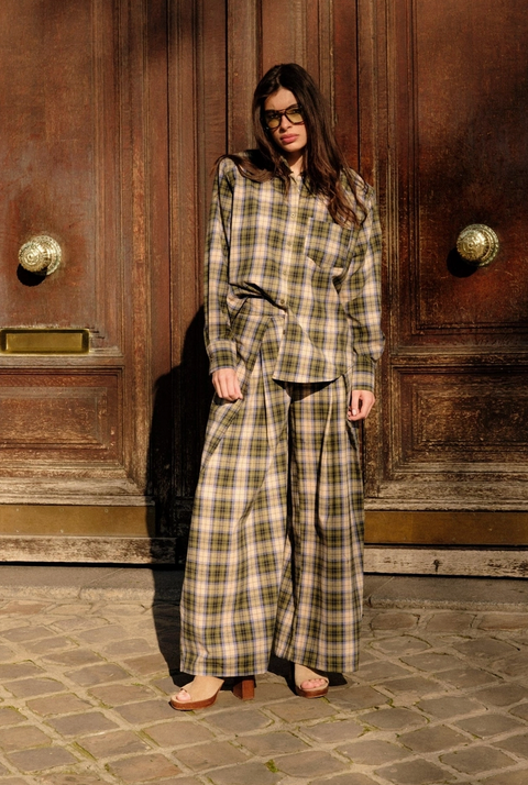 A woman wearing a matching plaid button-up shirt and wide-leg trousers stands in front of a large rustic wooden door on a cobblestone street.